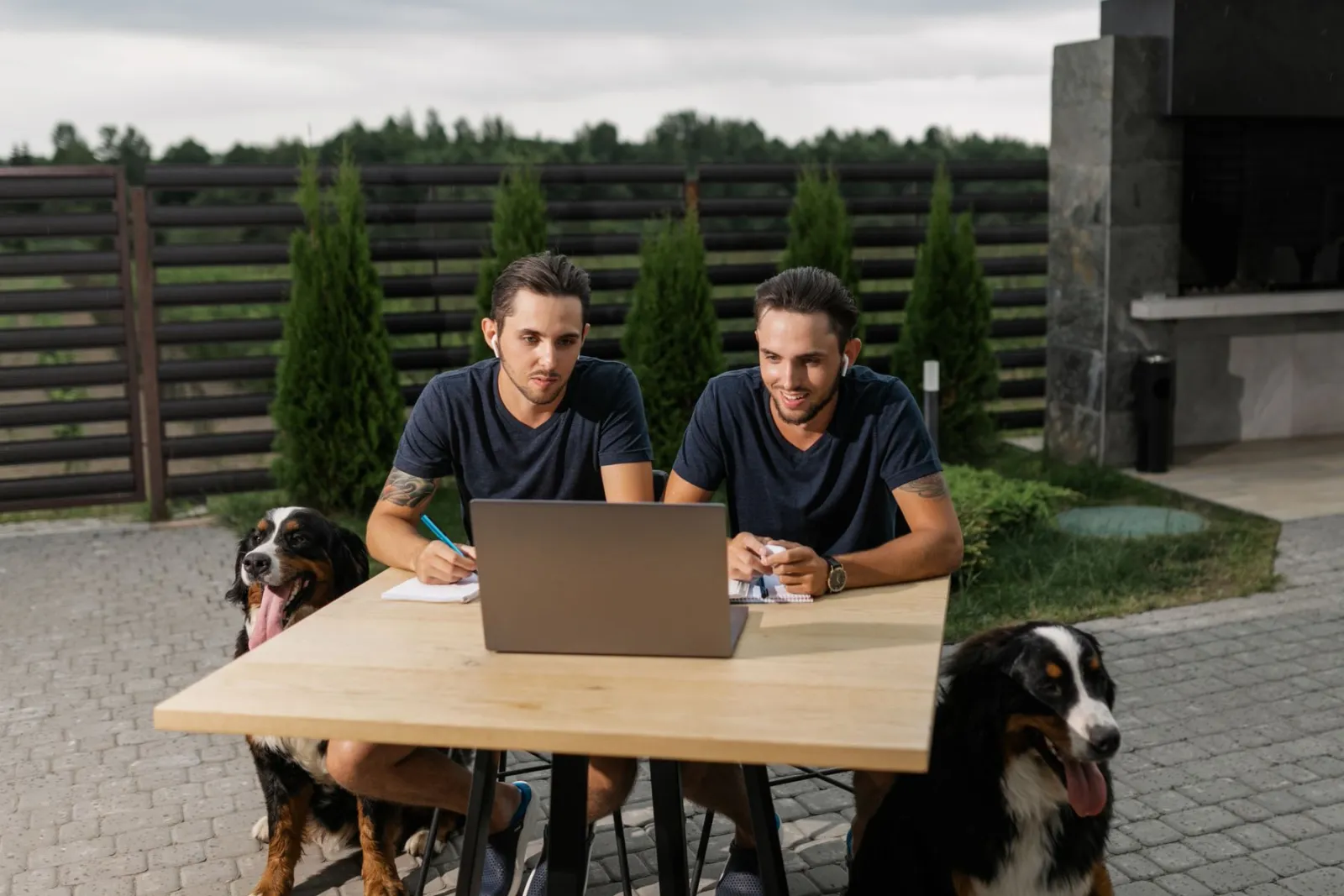 Dois tutores sentados diante de um notebook numa mesa ao ar livre, com seus dois cachorros ao lado